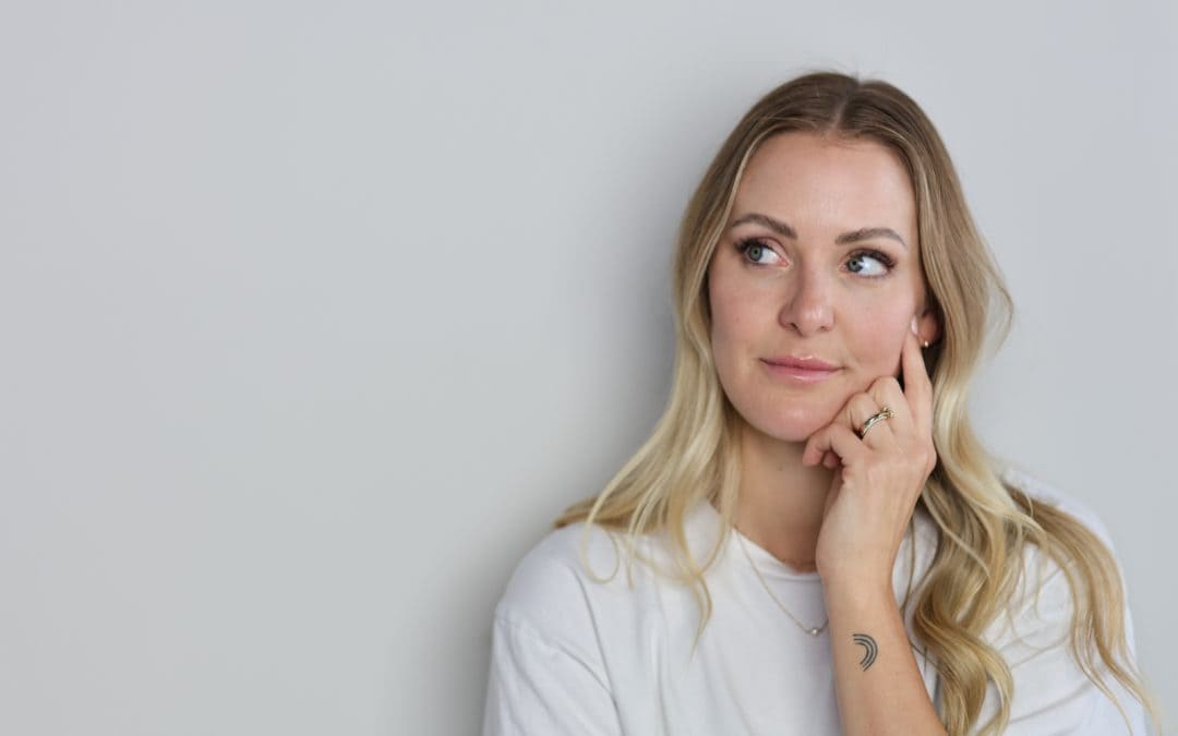Portrait of a blonde woman looking to her right, resting her chin on her left hand; crescent tattoo on her forearm visible.